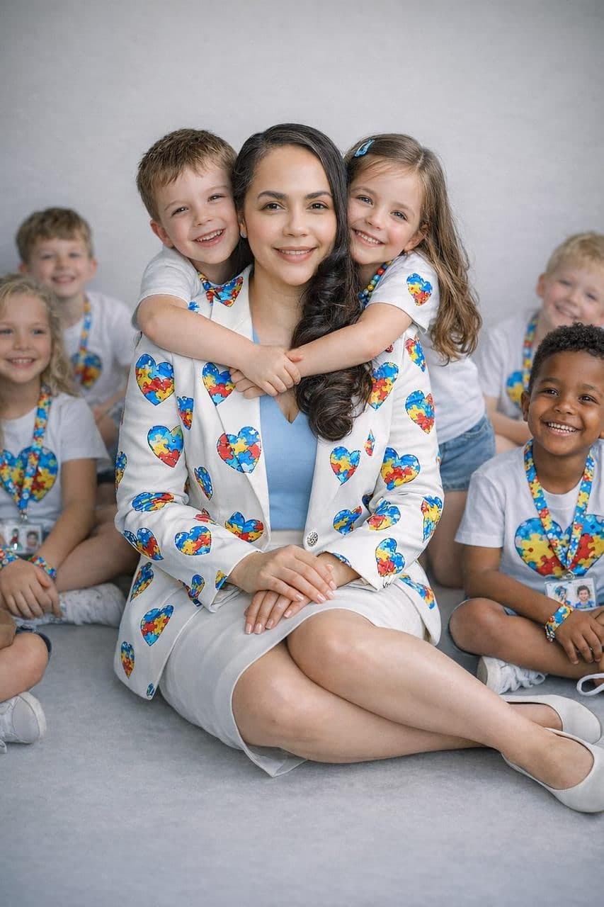 Advogada sentada no chão, abraçada por duas crianças, com outras crianças ao redor usando camisetas com o coração de quebra-cabeça da conscientização do autismo.